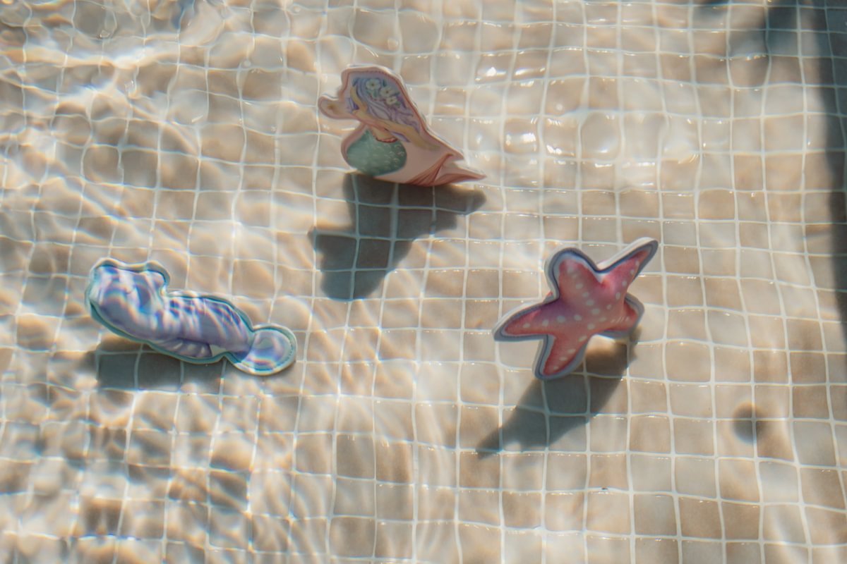 image_41632 Three cartoon-style paper cutouts of sea creatures lying on a tiled pool bottom: a pink starfish, a mermaid, and another blue-purple sea creature.
