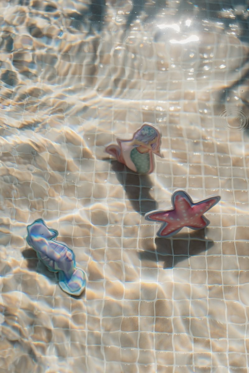image_41633 Small plastic pool toys (mermaid, pink starfish, blue seahorse) float on a sunlit pool bottom with a grid pattern.