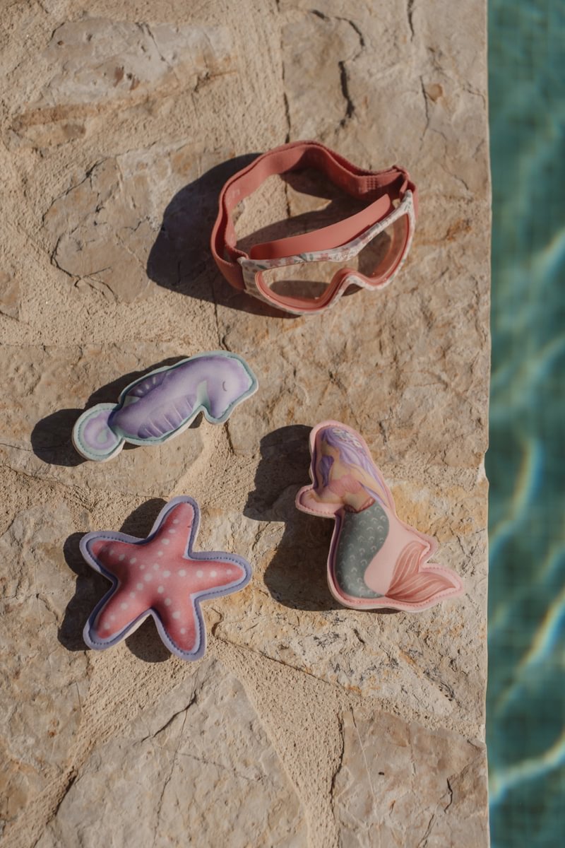 image_41635 Kids' swimming accessories laid on a stone pool edge: pink goggles, a purple seahorse toy, a pink starfish, and a pastel mermaid tail plush.