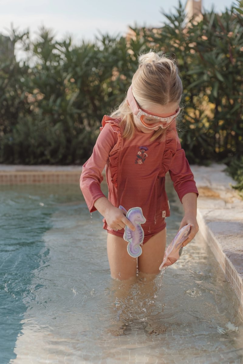 image_41636 Young girl in a pink swimsuit with goggles plays in a shallow pool, holding purple seahorse toys.