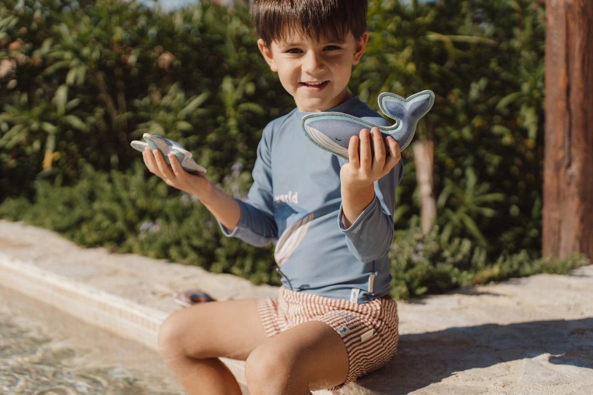 image_41649 Young boy sitting by a pool, holding a plush blue whale toy in one hand and cards in the other.