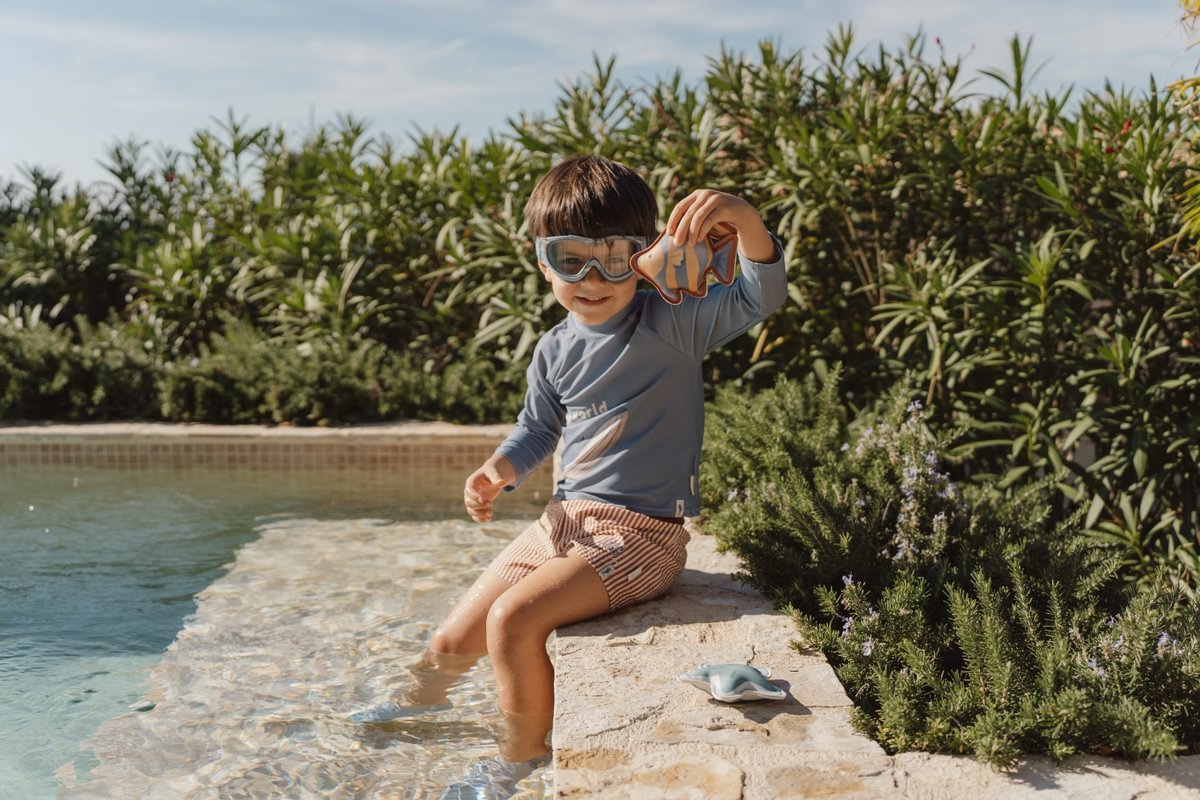 image_41651 Young child in a blue swim shirt and striped shorts sits at the pool edge with feet in the water, holding a small toy sea creature while wearing snorkeling goggles.