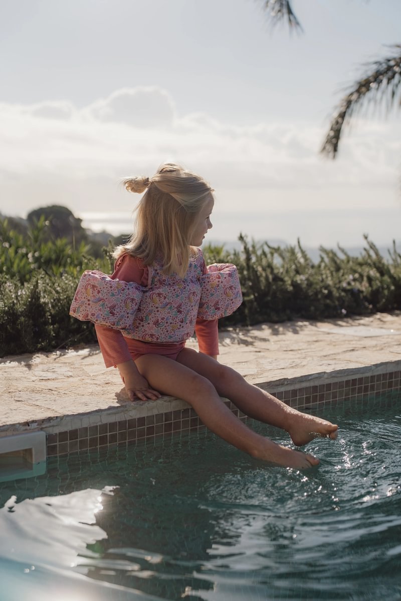 image_41672 Little girl in a pink floral swim top sits on the pool edge with her feet dipped in the water on a sunny day.