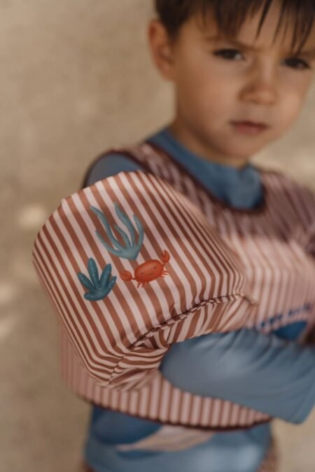 Close-up of a child wearing a pink striped jacket with blue sleeves, featuring a blue seaweed and orange crab patch on the sleeve.
