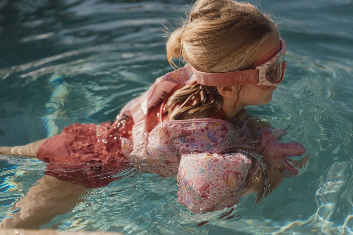 image_41675 Young girl swimming in a pink life jacket with unicorn-pattern arm floats and pink goggles in a blue pool.