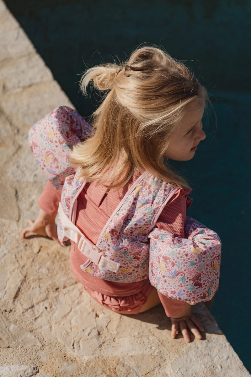 image_41676 Young girl sits on a stone ledge beside a blue pool, wearing a pink floral life jacket and pink swimsuit.