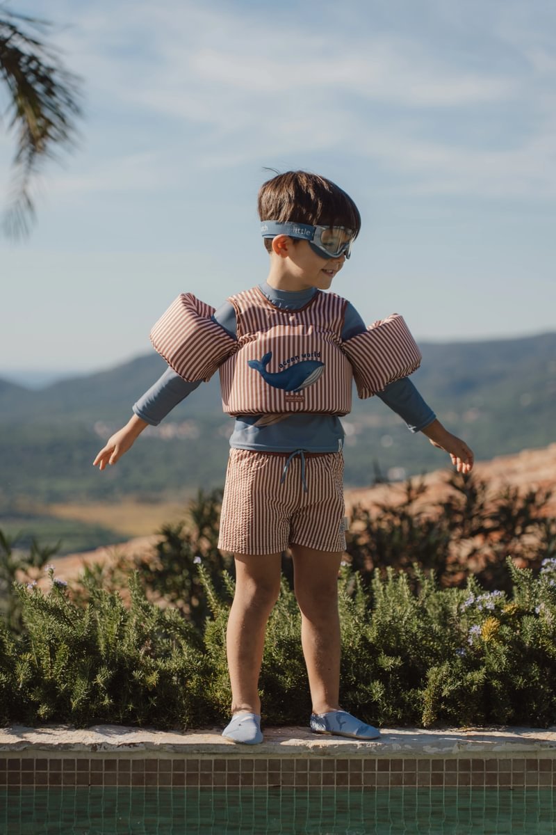 image_41680 Boy wearing a striped swim vest and blue swim shirt with goggles stands at the pool edge outdoors with mountains in the background