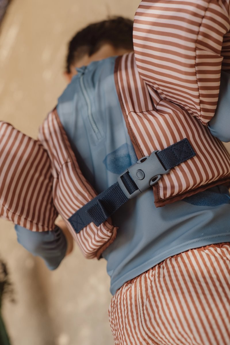 image_41681 Child wearing an orange-and-white striped life jacket with a blue interior, seen from behind with a gray buckle strap