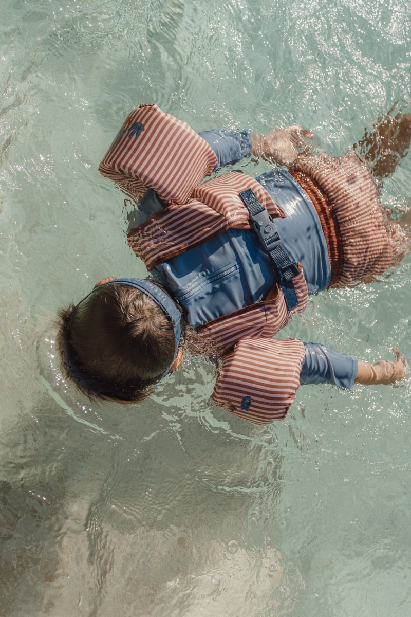 image_41682 Child wearing a blue life jacket and orange striped sleeves floating in clear shallow water, arms outstretched for balance.
