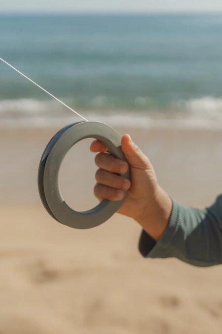 Person holding a circular grey ring toy with a string on a sandy beach, ocean in the background.
