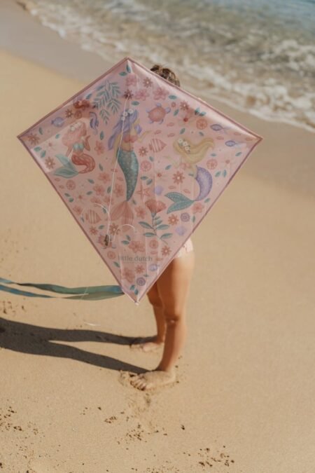 Child on a sunny beach holding a pink diamond-shaped kite with mermaids and sea creatures in the design at the shore.
