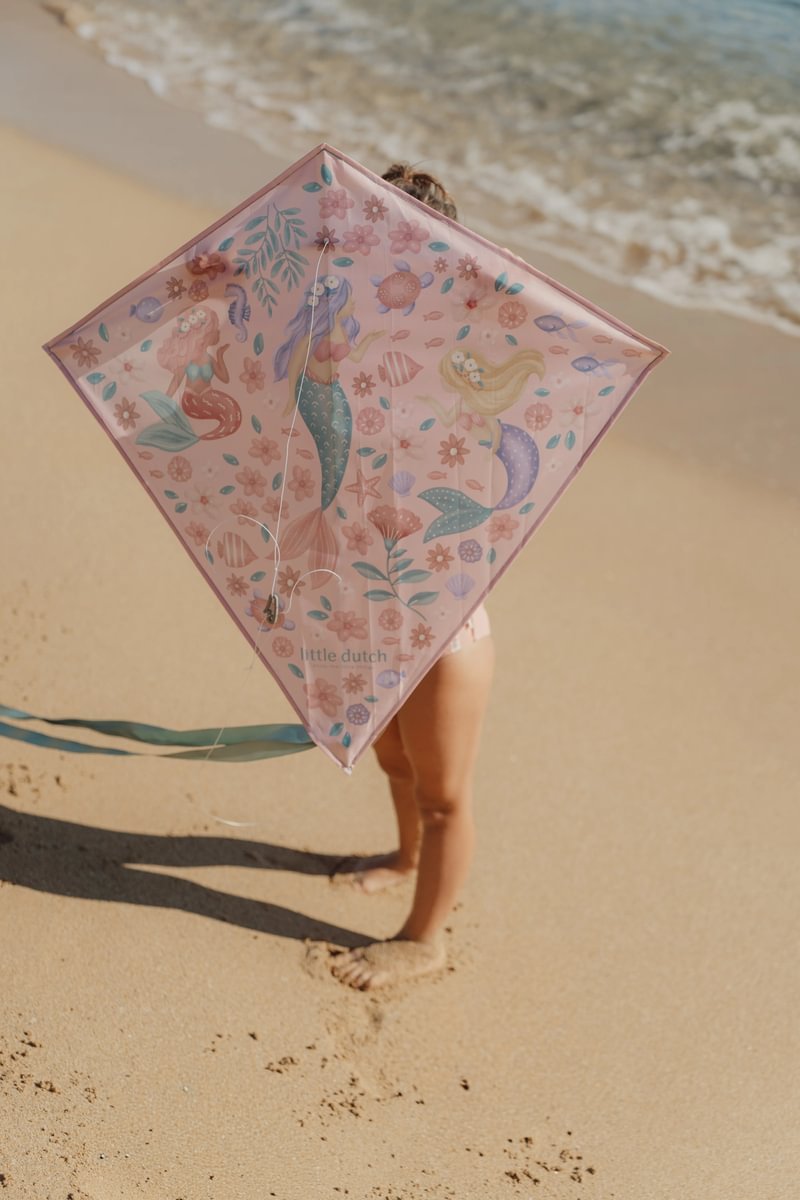 image_41699 Child on a sunny beach holding a pink diamond-shaped kite with mermaids and sea creatures in the design at the shore.