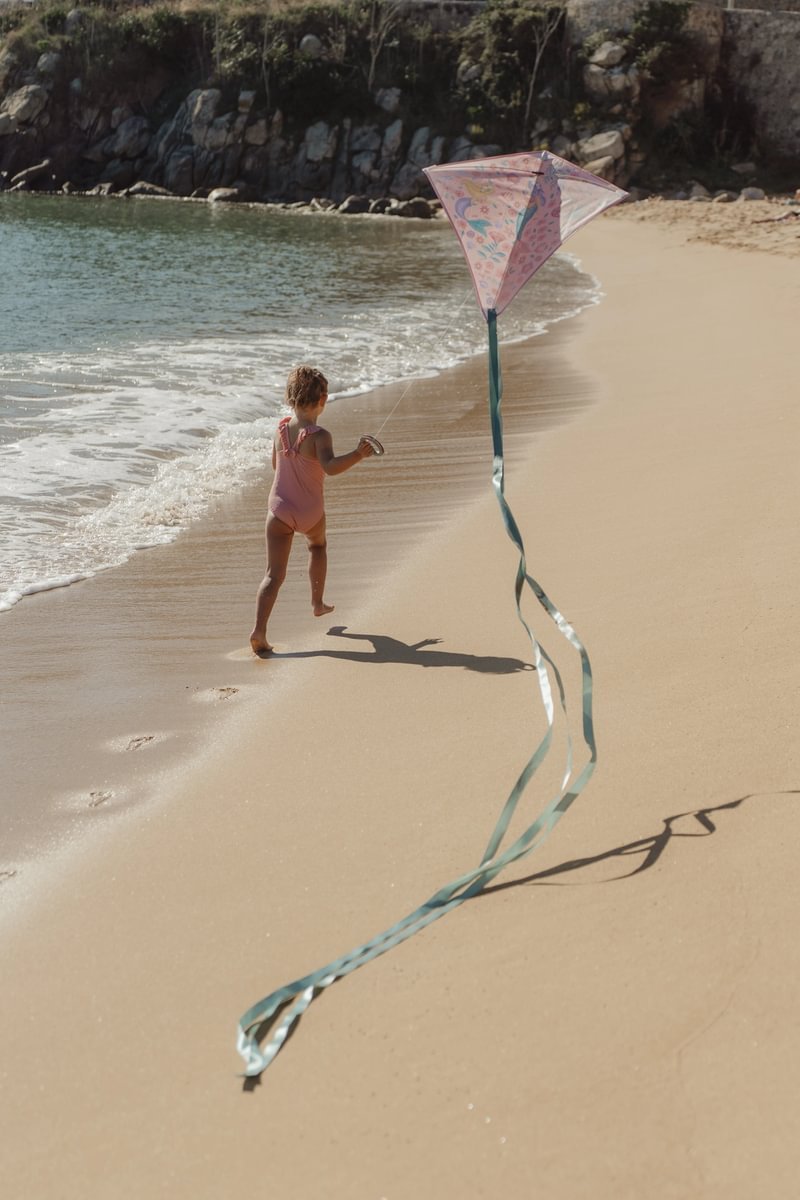 image_41700 Child in a pink swimsuit runs along a sandy beach with a pink kite and a long teal-tailed streamer.