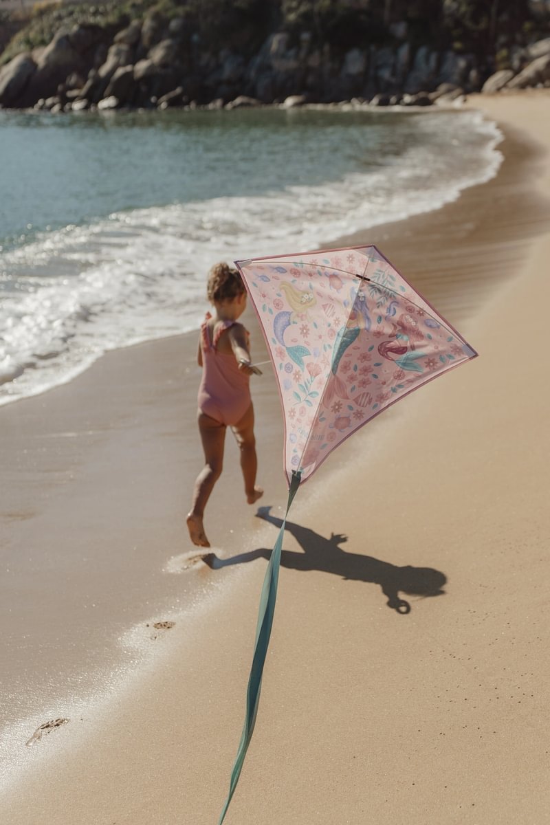 image_41701 Young child in a pink swimsuit runs along the sandy beach while holding a pink patterned kite on a string near the water's edge.
