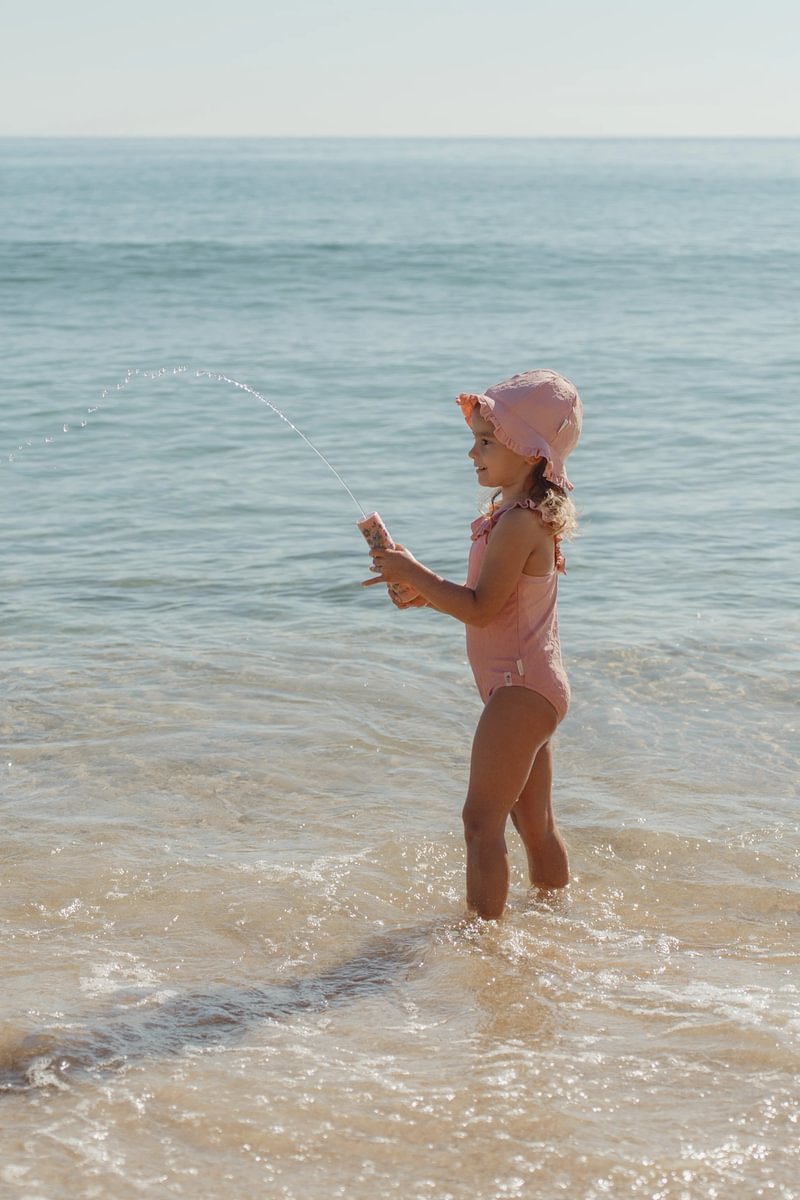 image_41769 Little girl in a pink swimsuit and sun hat plays with a water-spraying toy at the shoreline, standing in shallow surf.