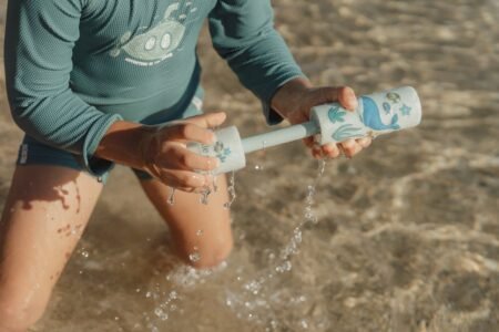 Child in a teal top grips a foam-ended water toy, spraying water as they play in shallow water.