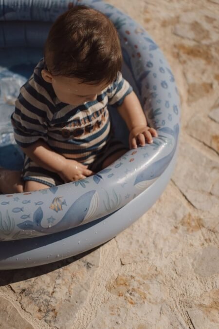 Toddler sits in a blue inflatable pool on a rocky beach, wearing a striped navy and beige outfit and looking down at hands.