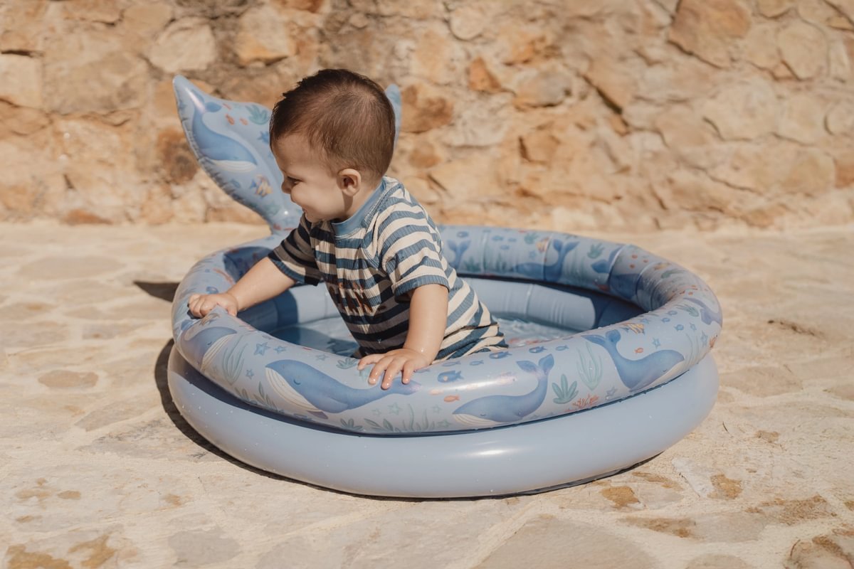 image_41780 Baby in a striped shirt sitting in a blue inflatable pool on a sunlit stone patio with a stone wall in the background.