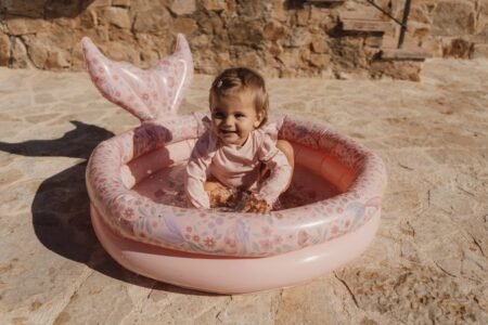 Baby in a pink outfit sits in a round pink inflatable kiddie pool outdoors, smiling as water splashes."No extra quotes"