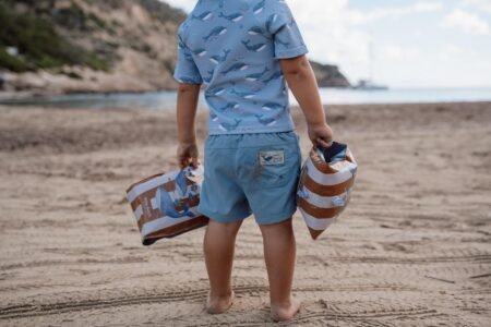 Child walking barefoot on a sandy beach, seen from behind, carrying two striped inflatable bags in each hand, wearing a blue whale-pattern shirt and shorts.