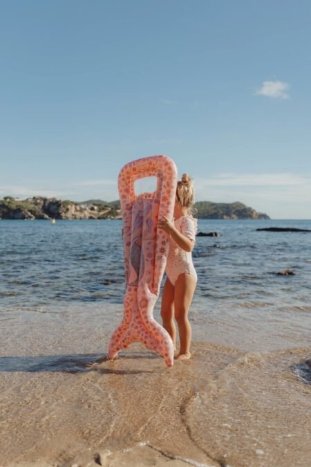 Child at the shore holding a large pink inflatable flamingo pool float on the beach.