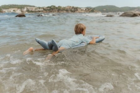Child lying on a light-blue bodyboard in shallow, sunlit sea with gentle waves at the beach.