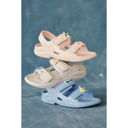 Three pastel sandals stacked from top to bottom (pink, white, blue) with small decorative charms on the straps against a blue background, including a leafy charm, a pineapple, and a tiny car.