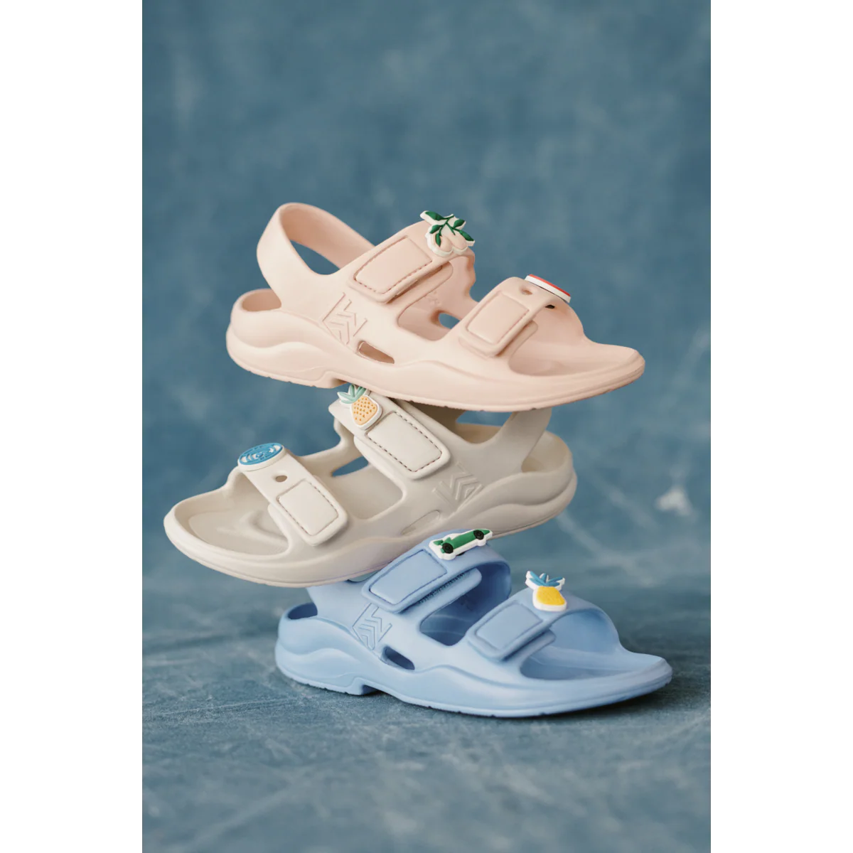 a29udGFpbmVyL25ldy80OTQ5LzE3ODc5NzM3djEucG5n Three pastel sandals stacked from top to bottom (pink, white, blue) with small decorative charms on the straps against a blue background, including a leafy charm, a pineapple, and a tiny car.