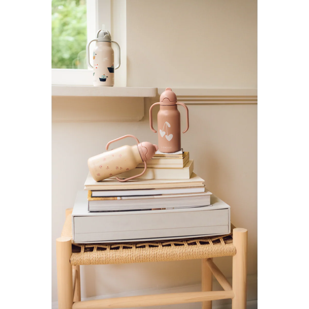 a29udGFpbmVyL25ldy80OTQ5LzE5MzAzMDUxdjEucG5n_5949efdf-8cbb-43fc-9058-ae729e2ee0d9 Pastel pink and cream bottles placed on a stack of books atop a woven stool in a cozy indoor scene.