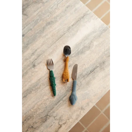Three kids’ utensils laid on a light marble countertop: green-handled fork, orange giraffe-spoon, and blue kids’ knife.