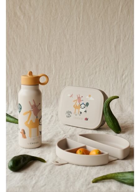 Kids' lunch set with playful animal illustrations (giraffes, panda) on a beige bottle and a square lunchbox, placed on a light fabric background with cucumber spears nearby.